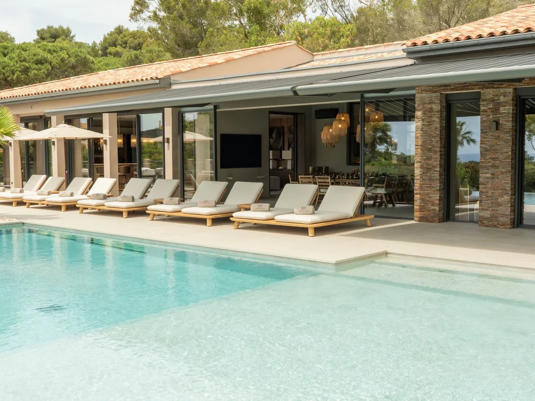 Grande piscine rectangulaire avec rangée de transats en teck et ouverture sur espace repas couvert